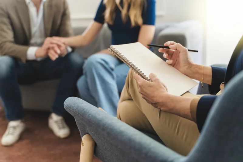 Couple Therapy Insight: Focus on Therapist’s Notebook with Young Married Couple in Background